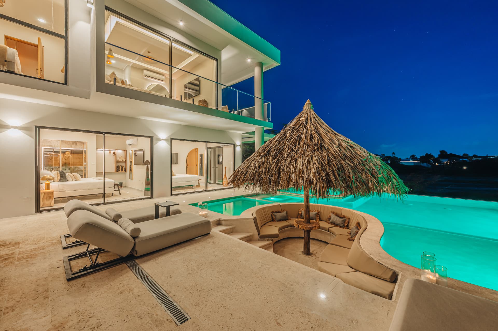 A Mediterranean-inspired villa at twilight, glowing with turquoise pool light and a thatched palapa